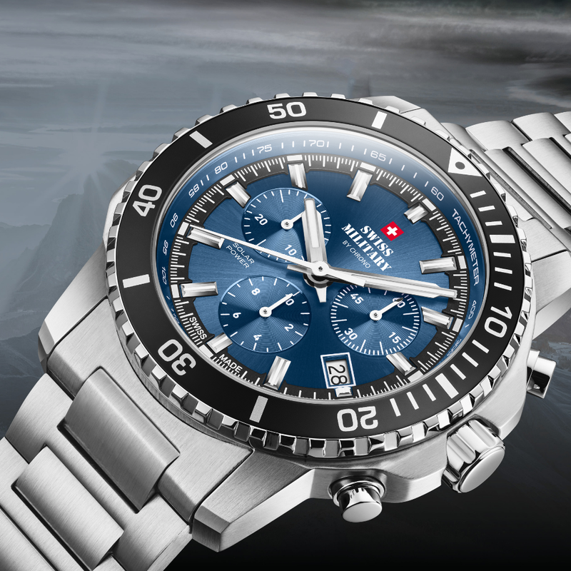 A close-up of the Swiss Military Solar Chronograph watch SMS34113 with a blue dial, black bezel, and silver metal bracelet, shown against a dark blurred background.