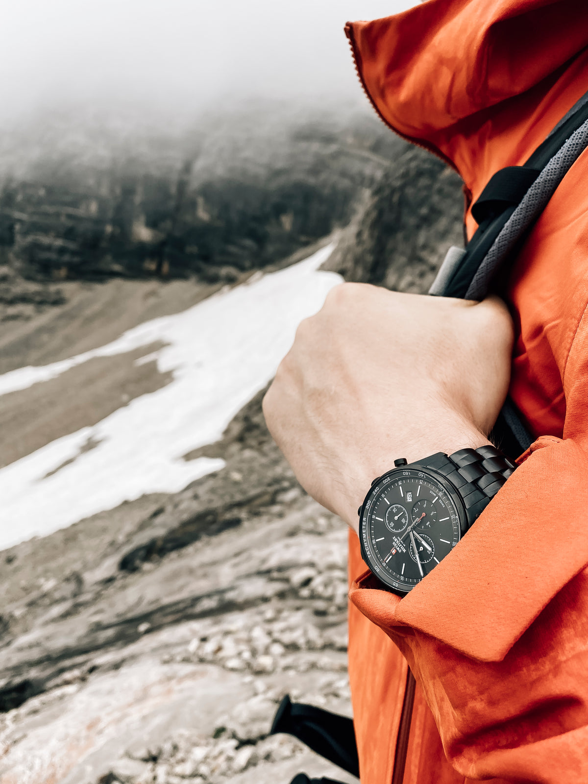 Person wearing an orange jacket and black watch in a mountainous landscape with snow.