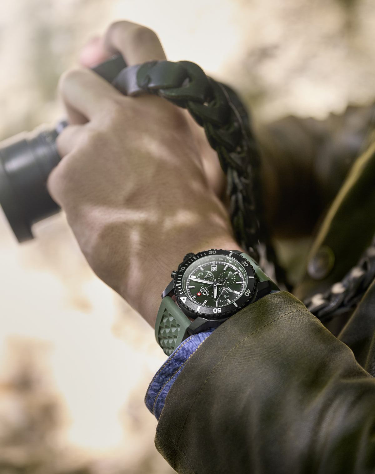 A close-up lifestyle shot of a person wearing a rugged green and black Swiss Military Solar watch, holding a camera with a blurred outdoor background.