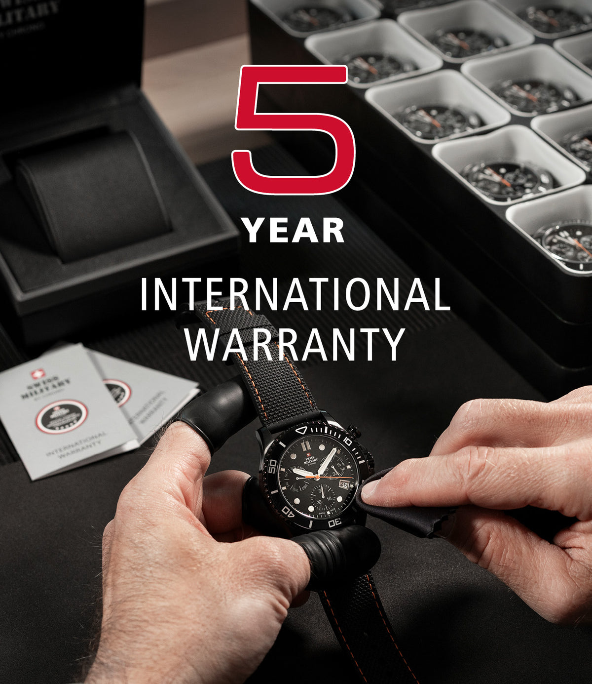A close-up of a Swiss Military watchmaker's hands holding a black wristwatch with a black strap and black dial, with a large red and white text overlay reading '5 YEAR INTERNATIONAL WARRANTY'. In the background, there are open black watch boxes containing multiple watches, and two warranty cards are visible on the left.